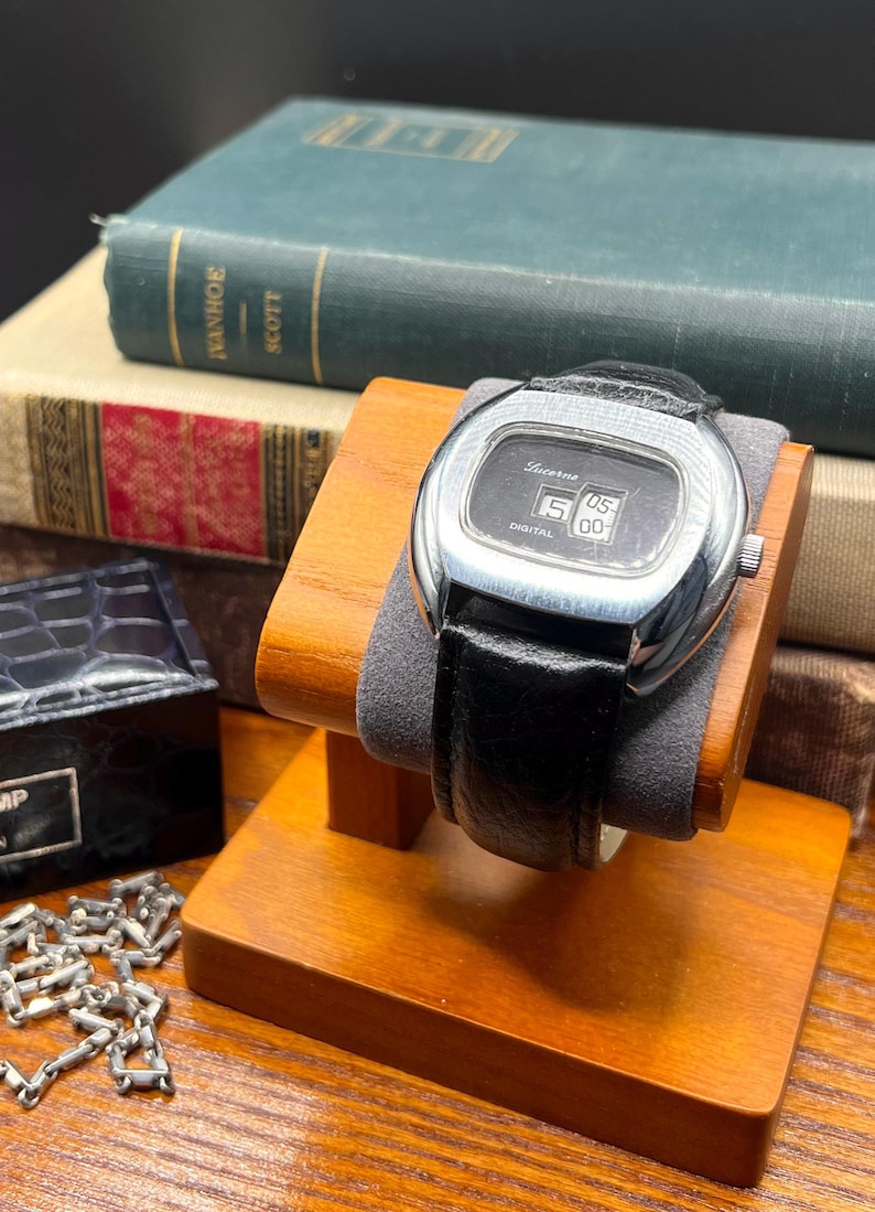 May include: A vintage Lucerne digital watch with a black leather band, displayed on a wooden stand. The watch face shows the time as 5:00. The watch has a silver-colored case and is set against a backdrop of old books.