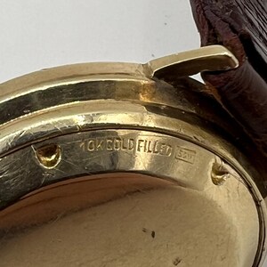 Puede incluir: Primer plano de una caja de reloj dorada con una correa de cuero marr&oacute;n. La caja del reloj tiene el texto "10K GOLD FILLED" y otras marcas. El reloj parece ser vintage.