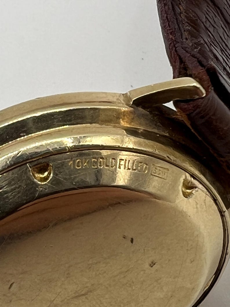 Puede incluir: Primer plano de una caja de reloj dorada con una correa de cuero marr&oacute;n. La caja del reloj tiene el texto "10K GOLD FILLED" y otras marcas. El reloj parece ser vintage.