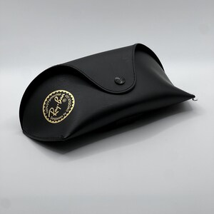 May include: Black sunglasses case with a snap closure. The case has a curved design and a gold emblem that reads "Ray-Ban Sunglasses by Luxottica". The case is made of a smooth material and is designed to protect eyewear.
