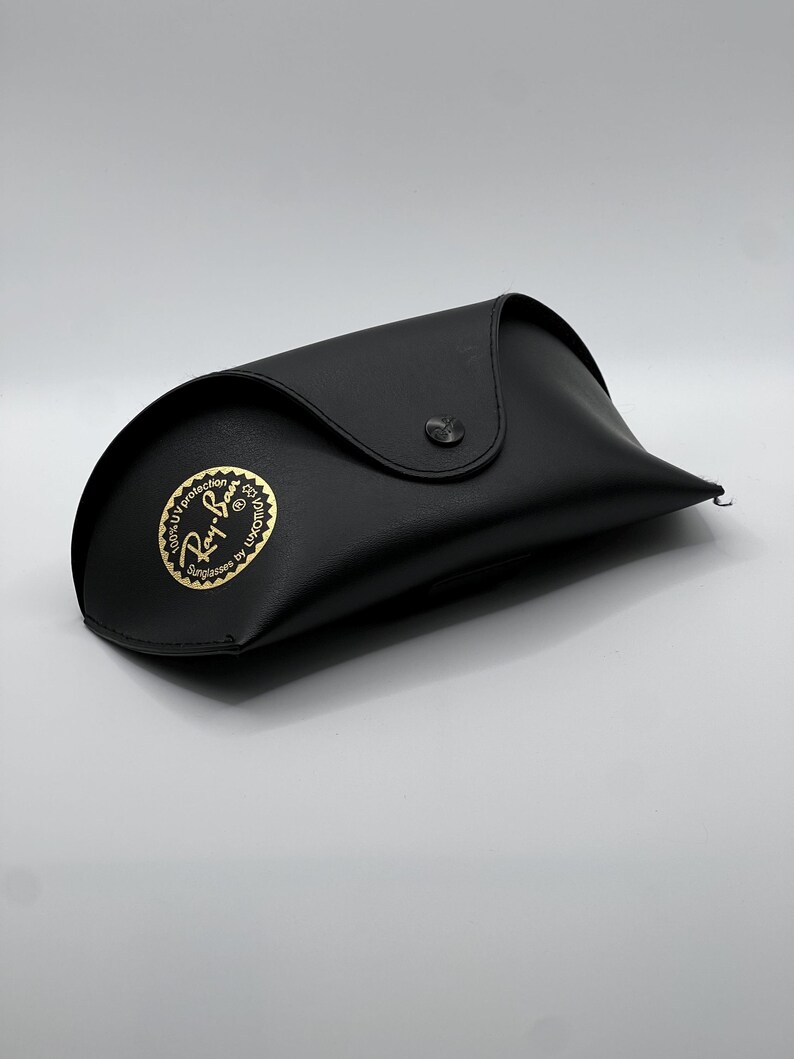 May include: Black sunglasses case with a snap closure. The case has a curved design and a gold emblem that reads "Ray-Ban Sunglasses by Luxottica". The case is made of a smooth material and is designed to protect eyewear.