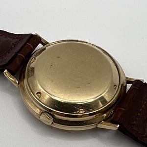 Puede incluir: Un reloj de pulsera dorado con una correa de cuero marr&oacute;n. La caja del reloj es redonda y tiene un acabado pulido. La correa del reloj es de color marr&oacute;n oscuro y parece ser de cuero.