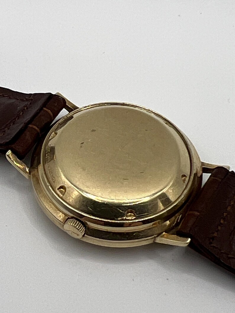 Puede incluir: Un reloj de pulsera dorado con una correa de cuero marr&oacute;n. La caja del reloj es redonda y tiene un acabado pulido. La correa del reloj es de color marr&oacute;n oscuro y parece ser de cuero.