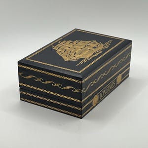 Puede incluir: Caja rectangular negra con detalles dorados y una ilustraci&oacute;n dorada de un barco en la tapa. La palabra "LONGINES" est&aacute; impresa en dorado en el lateral. Para joyas o un reloj.