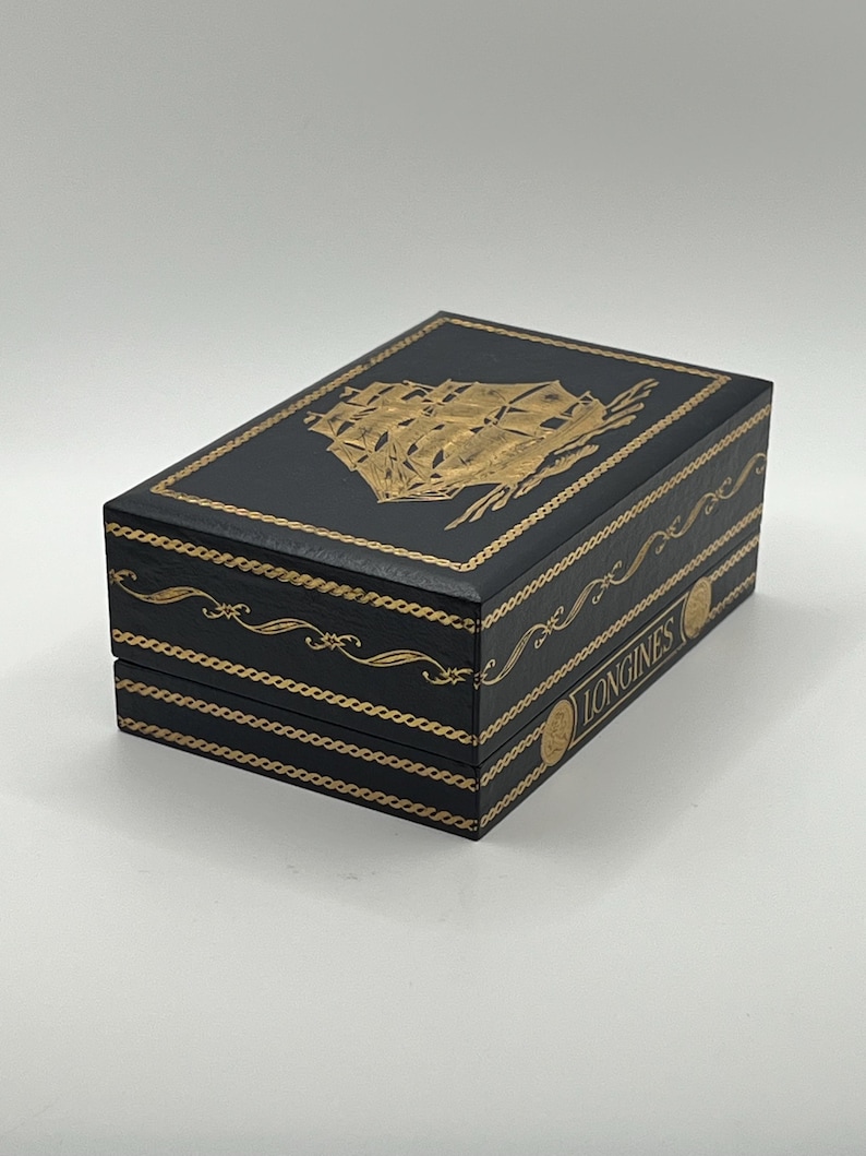 Puede incluir: Caja rectangular negra con detalles dorados y una ilustraci&oacute;n dorada de un barco en la tapa. La palabra "LONGINES" est&aacute; impresa en dorado en el lateral. Para joyas o un reloj.