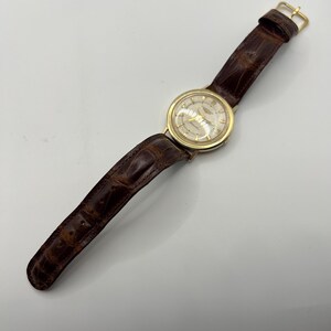Puede incluir: Un reloj de pulsera dorado con una correa de cuero marr&oacute;n. La esfera del reloj es de color claro con agujas y marcadores dorados. La correa es de color marr&oacute;n oscuro con un patr&oacute;n texturizado similar al de un cocodrilo. El reloj es de estilo cl&aacute;sico.