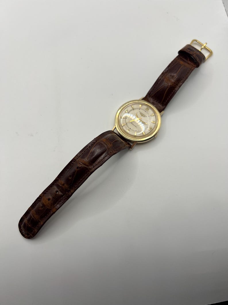 Puede incluir: Un reloj de pulsera dorado con una correa de cuero marr&oacute;n. La esfera del reloj es de color claro con agujas y marcadores dorados. La correa es de color marr&oacute;n oscuro con un patr&oacute;n texturizado similar al de un cocodrilo. El reloj es de estilo cl&aacute;sico.