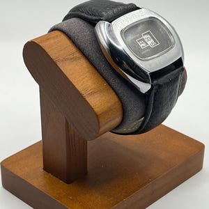 May include: A vintage digital watch with a black leather band and a silver-toned square face. The watch displays the time and is resting on a wooden watch stand. The stand is made of a light brown wood.