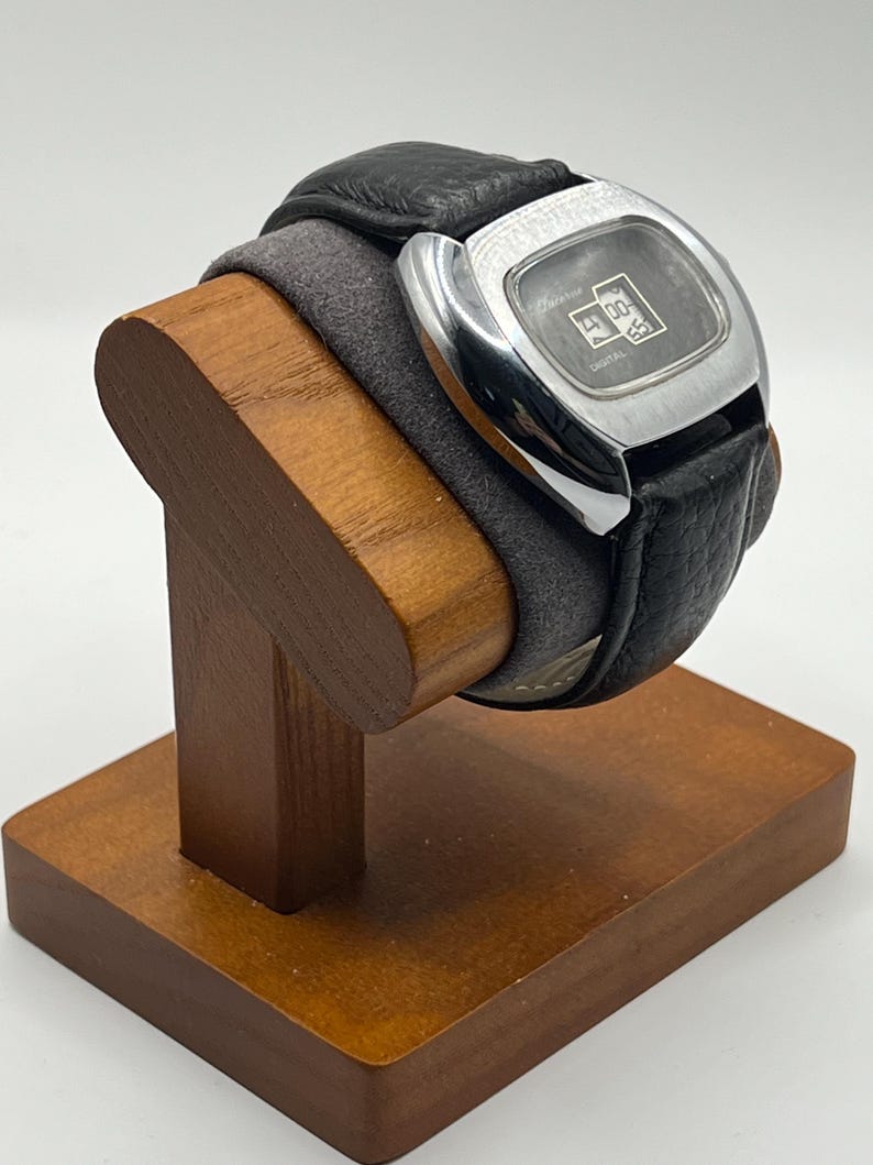 May include: A vintage digital watch with a black leather band and a silver-toned square face. The watch displays the time and is resting on a wooden watch stand. The stand is made of a light brown wood.
