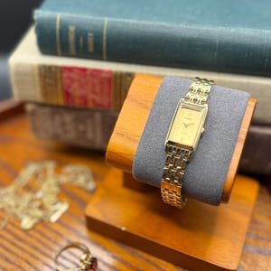 May include: A gold-toned rectangular wristwatch with a matching link bracelet rests on a wooden watch stand with a gray cushion. A gold ring with a red stone and a gold necklace are also visible.