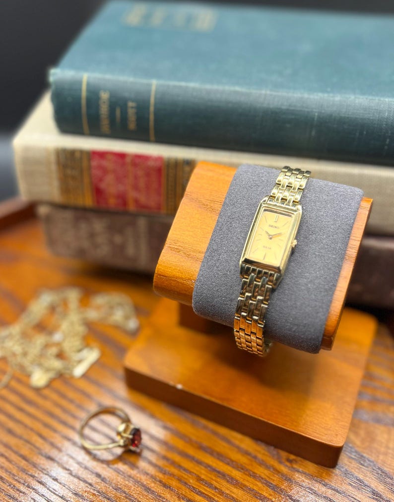 May include: A gold-toned rectangular wristwatch with a matching link bracelet rests on a wooden watch stand with a gray cushion. A gold ring with a red stone and a gold necklace are also visible.