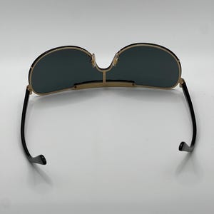 Peut inclure: Lunettes de soleil &agrave; monture dor&eacute;e avec verres vert fonc&eacute;. Les lunettes ont un double pont et des branches noires. Les verres sont de forme arrondie. Les lunettes sont pos&eacute;es sur un fond blanc.
