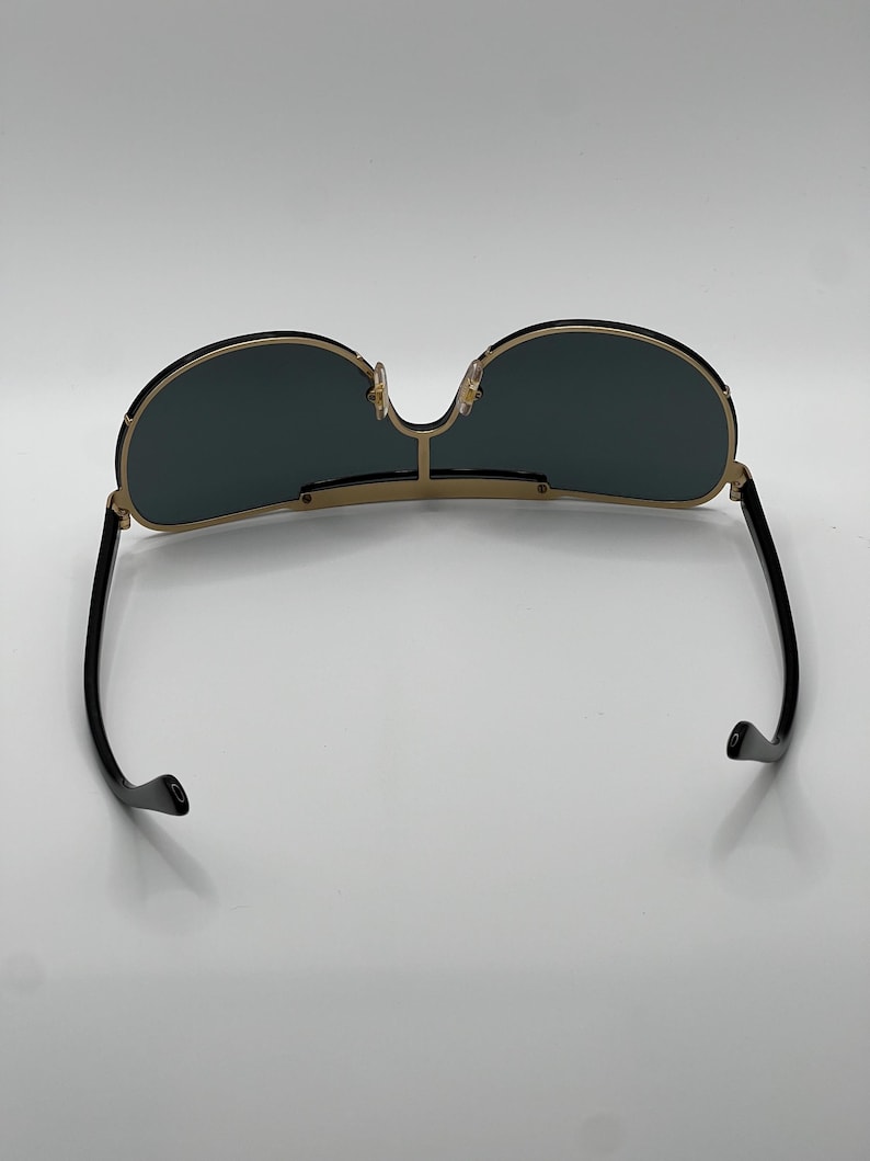 Peut inclure: Lunettes de soleil &agrave; monture dor&eacute;e avec verres vert fonc&eacute;. Les lunettes ont un double pont et des branches noires. Les verres sont de forme arrondie. Les lunettes sont pos&eacute;es sur un fond blanc.