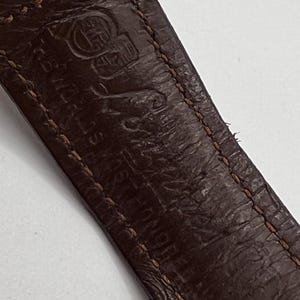 Puede incluir: Primer plano de una correa de reloj de cuero marr&oacute;n con costuras visibles y texto en relieve. El cuero tiene un aspecto texturizado y las costuras son de color marr&oacute;n claro. La correa muestra signos de desgaste.