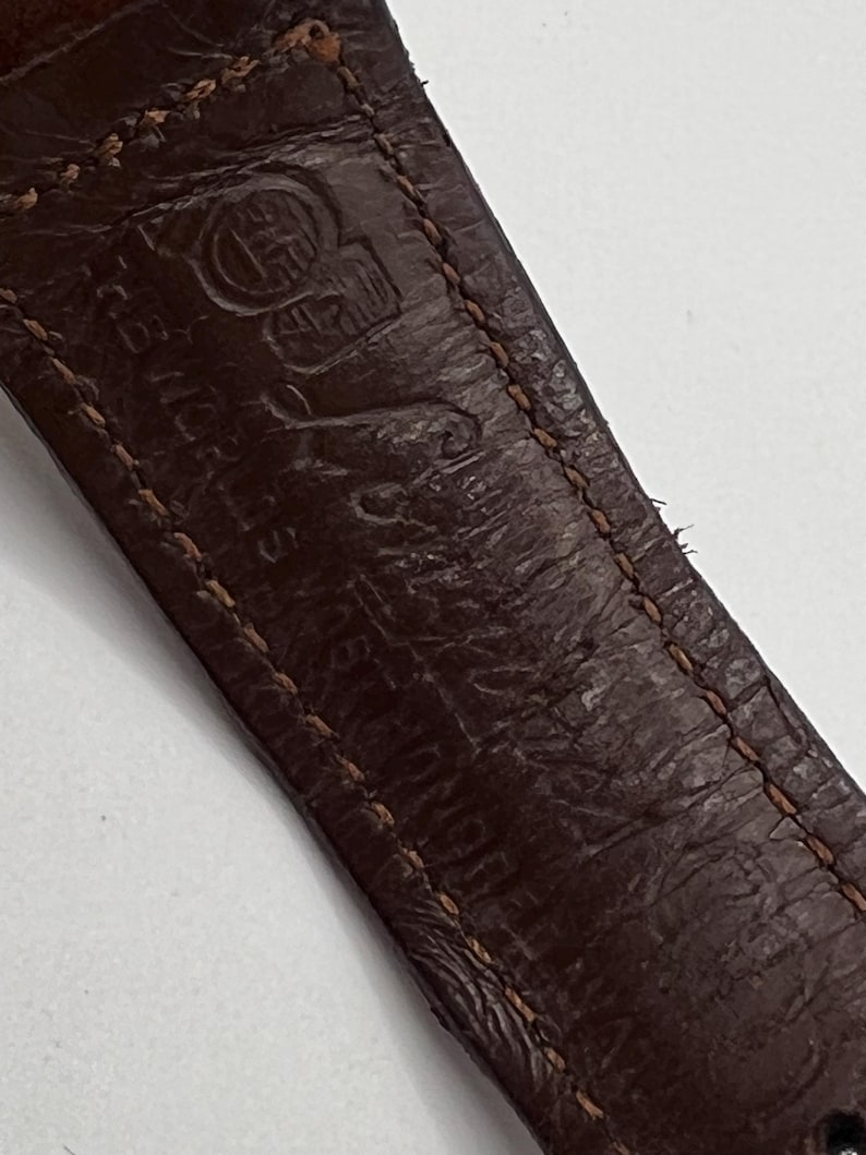 Puede incluir: Primer plano de una correa de reloj de cuero marr&oacute;n con costuras visibles y texto en relieve. El cuero tiene un aspecto texturizado y las costuras son de color marr&oacute;n claro. La correa muestra signos de desgaste.