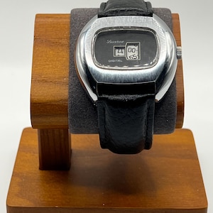 May include: A vintage Lucerne digital watch with a black leather band. The watch has a silver-colored rectangular case and a black face displaying the time. The watch is displayed on a wooden watch stand.