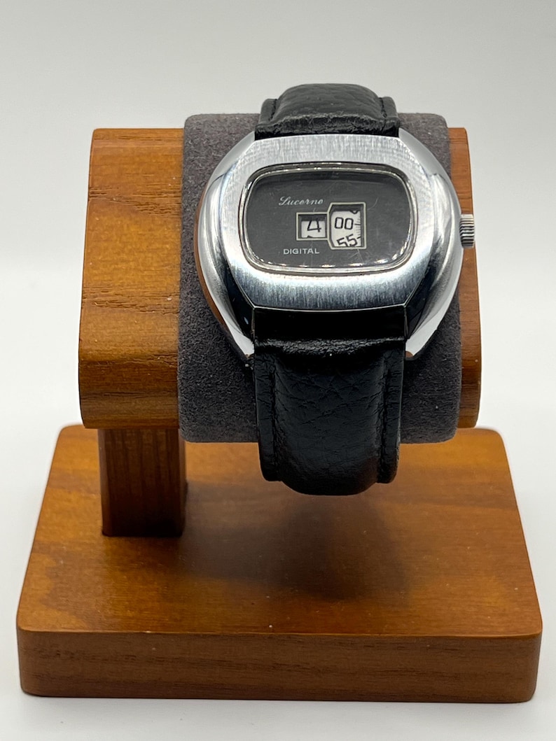 May include: A vintage Lucerne digital watch with a black leather band. The watch has a silver-colored rectangular case and a black face displaying the time. The watch is displayed on a wooden watch stand.