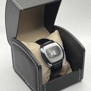 May include: A vintage silver-toned watch with a black leather band rests on a beige pillow inside a black leather watch box. The watch face displays the time. The box is rectangular with white stitching.