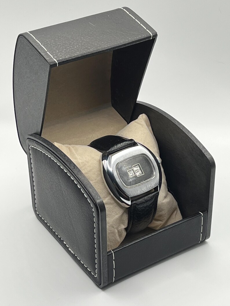 May include: A vintage silver-toned watch with a black leather band rests on a beige pillow inside a black leather watch box. The watch face displays the time. The box is rectangular with white stitching.