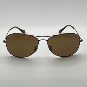 May include: Brown aviator sunglasses with a dark metal frame. The lenses are a solid brown color, and the frame has a classic teardrop shape. The sunglasses have the "Ray-Ban P" logo on the left lens.