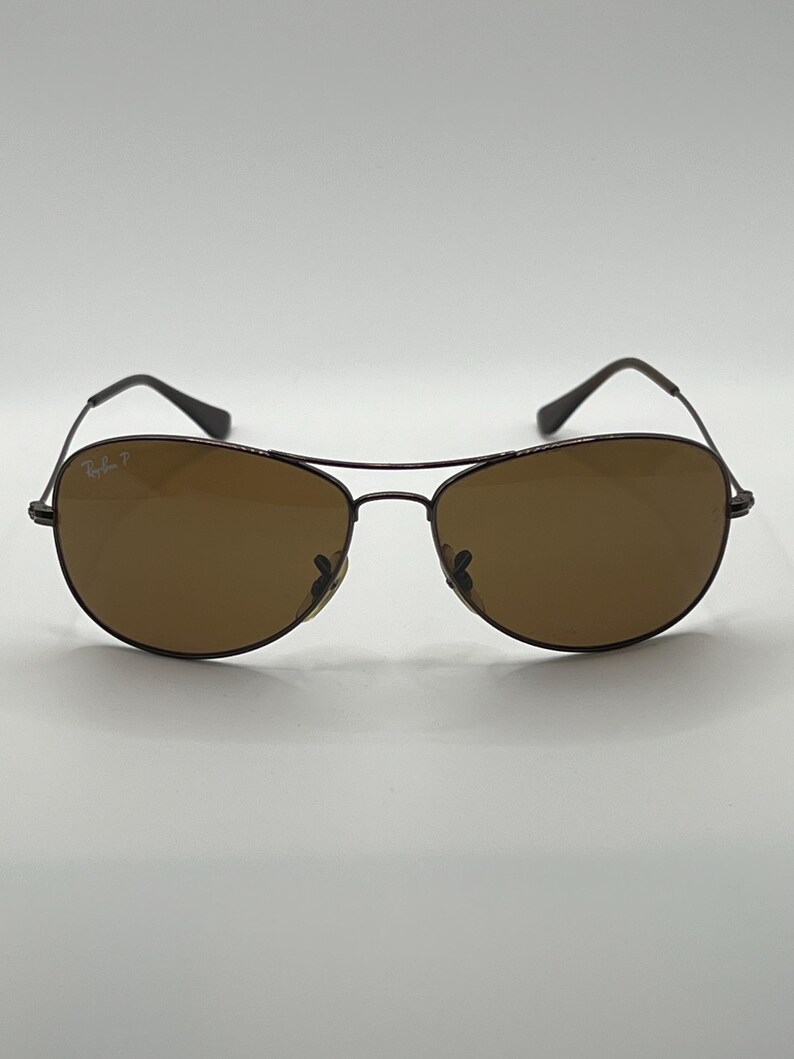 May include: Brown aviator sunglasses with a dark metal frame. The lenses are a solid brown color, and the frame has a classic teardrop shape. The sunglasses have the "Ray-Ban P" logo on the left lens.
