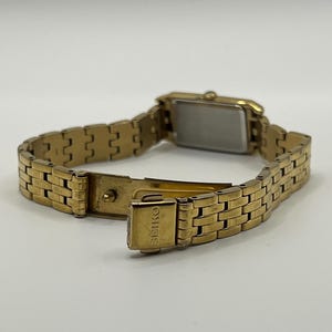 May include: A gold-toned Seiko wristwatch with a rectangular face and a linked bracelet. The watch is open, revealing the clasp and the Seiko brand name. The watch is set against a plain background.