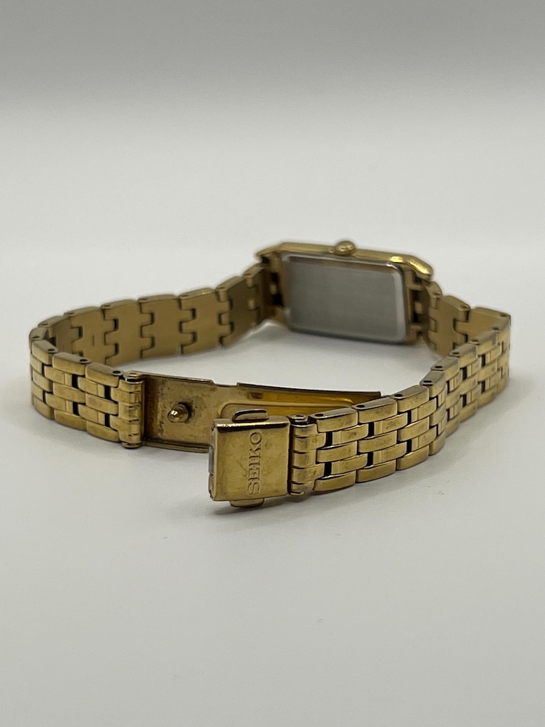 May include: A gold-toned Seiko wristwatch with a rectangular face and a linked bracelet. The watch is open, revealing the clasp and the Seiko brand name. The watch is set against a plain background.