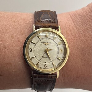 Puede incluir: Un reloj de pulsera autom&aacute;tico Longines dorado con esfera crema y correa de cuero marr&oacute;n. La esfera del reloj presenta agujas e indicadores dorados, con el logotipo de Longines y el texto "Automatic". La correa es de cuero marr&oacute;n oscuro texturizado.