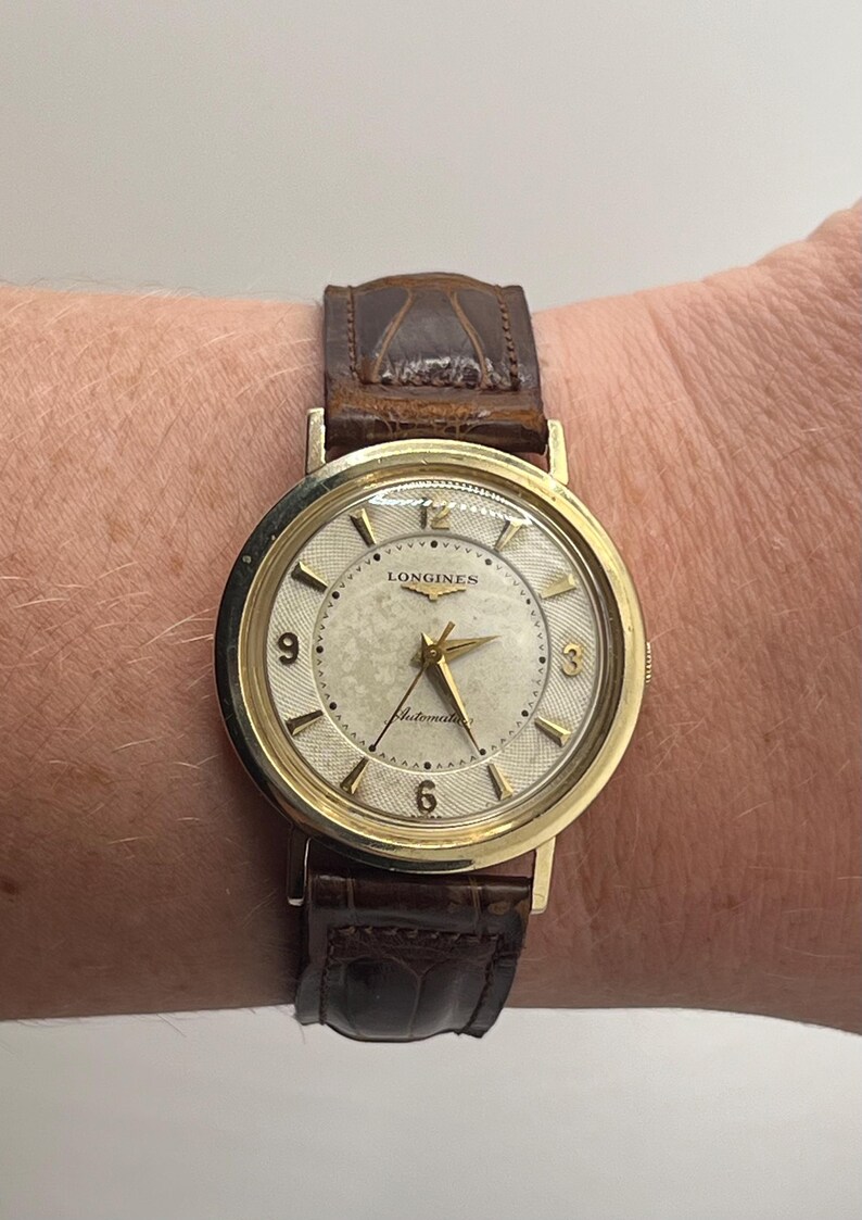 Puede incluir: Un reloj de pulsera autom&aacute;tico Longines dorado con esfera crema y correa de cuero marr&oacute;n. La esfera del reloj presenta agujas e indicadores dorados, con el logotipo de Longines y el texto "Automatic". La correa es de cuero marr&oacute;n oscuro texturizado.