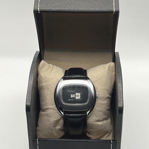 May include: A vintage digital watch with a black leather band, displayed in a black box with a cream-colored pillow. The watch face is silver with the word "Lucerne" and the time "15:00" displayed. The word "DIGITAL" is also visible.