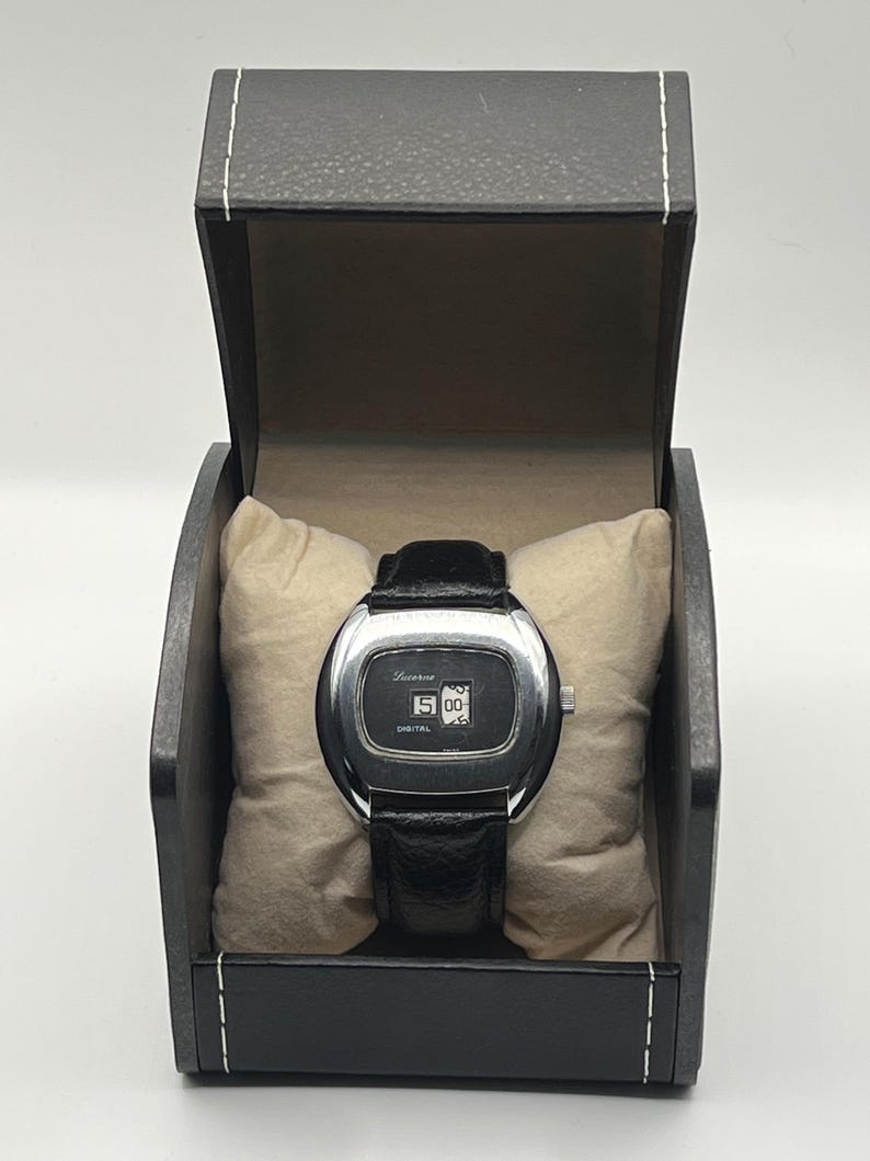 May include: A vintage digital watch with a black leather band, displayed in a black box with a cream-colored pillow. The watch face is silver with the word "Lucerne" and the time "15:00" displayed. The word "DIGITAL" is also visible.