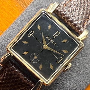 May include: A vintage Benrus wristwatch with a square gold-tone case and a black dial. The dial features gold-tone hour markers and hands, with the brand name "BENRUS" displayed. The watch has a brown leather strap.