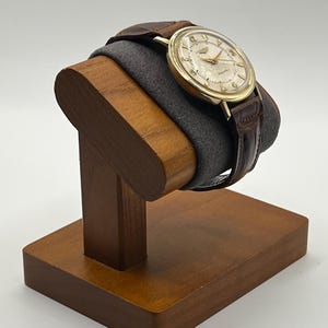 Puede incluir: Un reloj de pulsera vintage dorado con una correa de cuero marr&oacute;n se exhibe en un soporte de reloj de madera. El reloj tiene una esfera de color claro con manecillas y marcadores dorados. El soporte es de madera oscura y tiene un coj&iacute;n gris.
