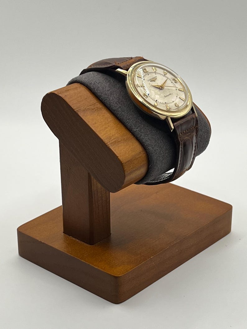 Puede incluir: Un reloj de pulsera vintage dorado con una correa de cuero marr&oacute;n se exhibe en un soporte de reloj de madera. El reloj tiene una esfera de color claro con manecillas y marcadores dorados. El soporte es de madera oscura y tiene un coj&iacute;n gris.