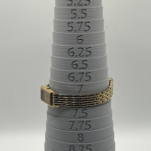 May include: A gold-toned rectangular watch with a linked band is placed on a gray ring sizer. The sizer has measurements from 5 to 8.75. The watch is set to a size 7.