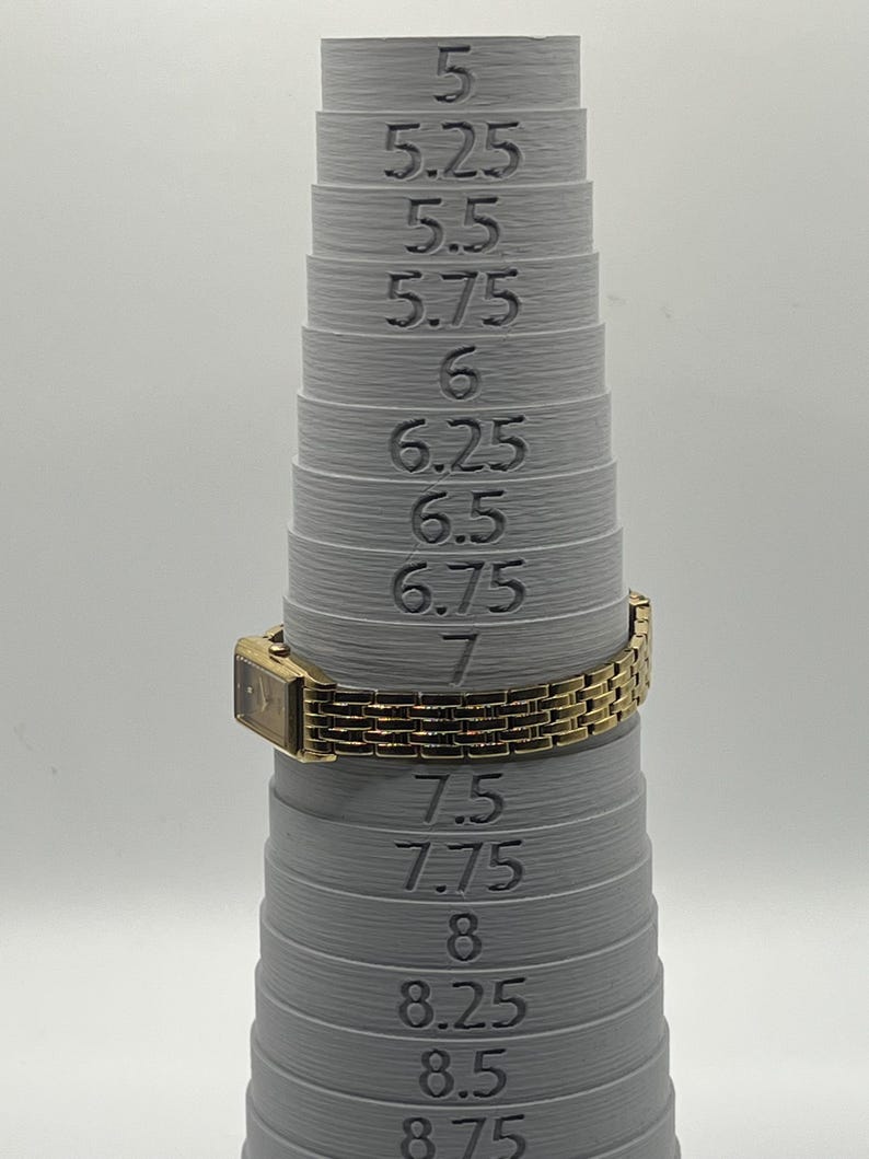 May include: A gold-toned rectangular watch with a linked band is placed on a gray ring sizer. The sizer has measurements from 5 to 8.75. The watch is set to a size 7.