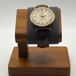Puede incluir: Un reloj de pulsera dorado vintage con una correa de cuero marr&oacute;n se exhibe en un soporte de madera para relojes. La esfera del reloj tiene un fondo crema con manecillas y n&uacute;meros dorados. El reloj est&aacute; marcado "Longines" y "Automatic".