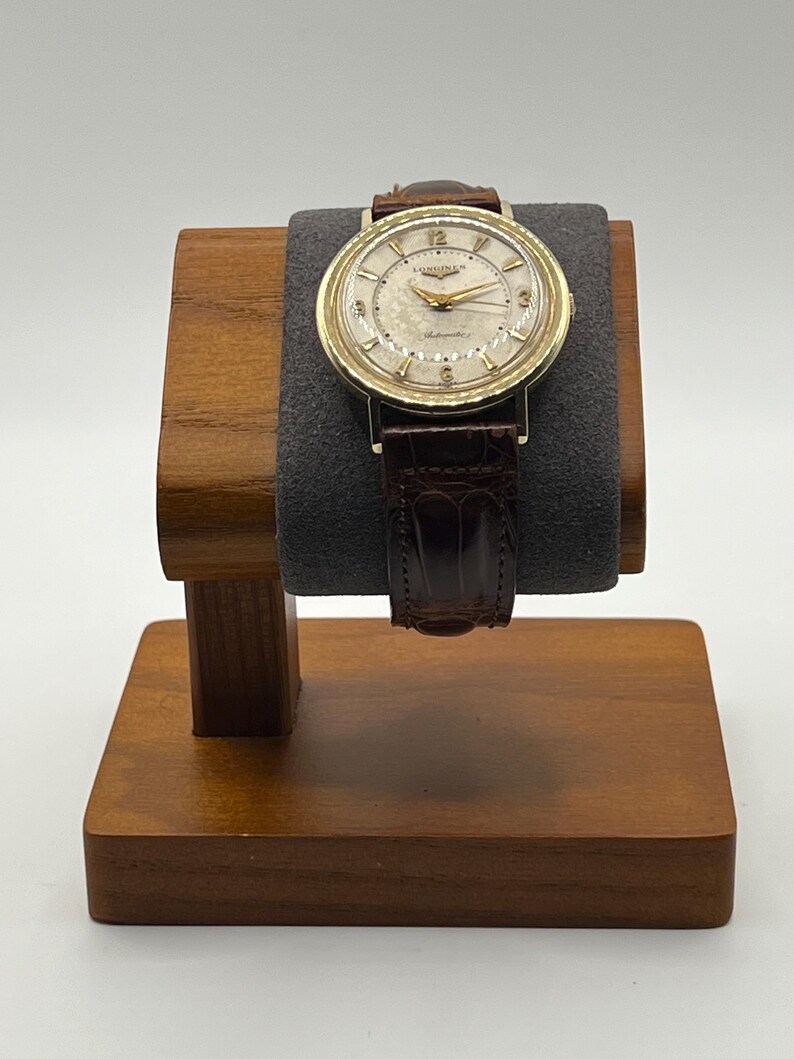 Puede incluir: Un reloj de pulsera dorado vintage con una correa de cuero marr&oacute;n se exhibe en un soporte de madera para relojes. La esfera del reloj tiene un fondo crema con manecillas y n&uacute;meros dorados. El reloj est&aacute; marcado "Longines" y "Automatic".