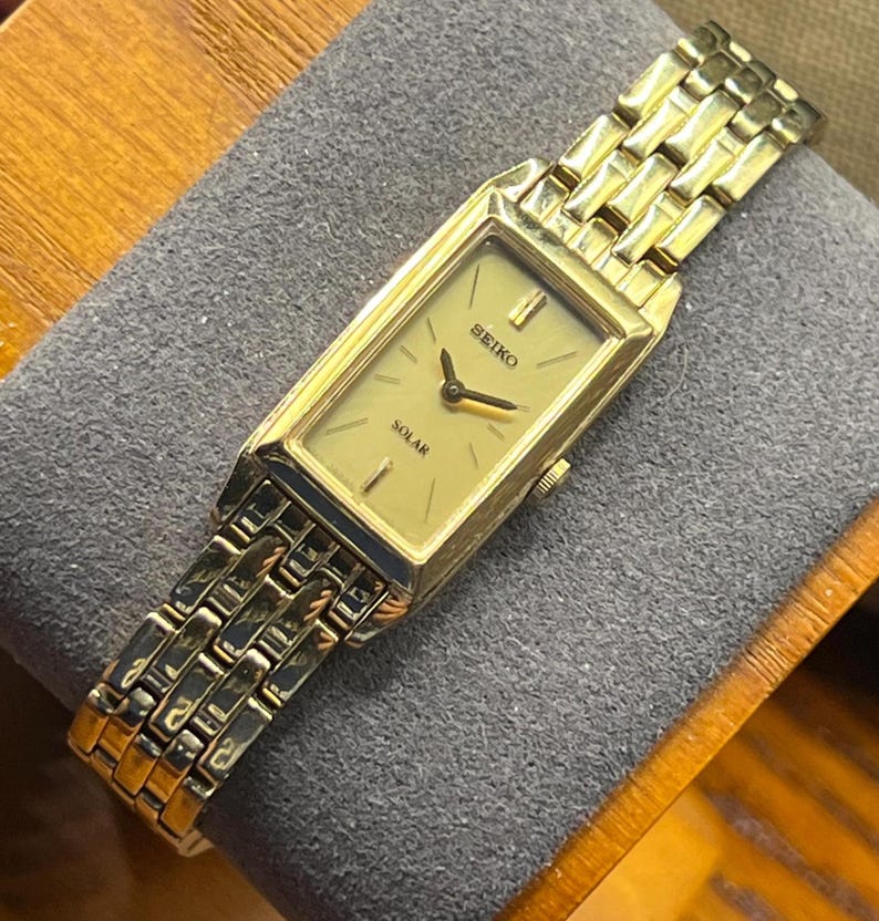 May include: A gold-toned Seiko Solar watch with a rectangular face and a matching gold link band. The watch face is a light gold color with black hands and the word "SEIKO" and "SOLAR" printed on it. The watch is displayed on a gray surface.