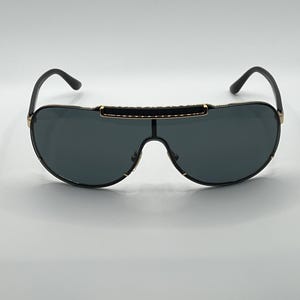 Peut inclure: Lunettes de soleil noires avec des verres gris fonc&eacute;. Les lunettes ont une monture noire avec des accents dor&eacute;s et une barre noire sur le dessus. Les branches sont noires et se courbent vers les oreilles.