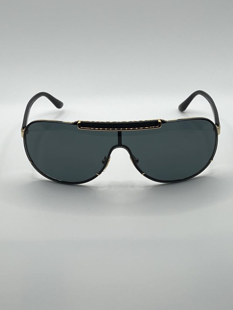 Peut inclure: Lunettes de soleil noires avec des verres gris fonc&eacute;. Les lunettes ont une monture noire avec des accents dor&eacute;s et une barre noire sur le dessus. Les branches sont noires et se courbent vers les oreilles.
