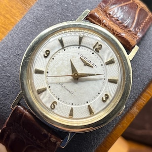 Puede incluir: Un reloj de pulsera autom&aacute;tico Longines vintage con esfera plateada, agujas e indicadores dorados y correa de cuero marr&oacute;n. La esfera del reloj muestra el logotipo de Longines y la palabra "Automatic". El reloj tiene un bisel dorado.