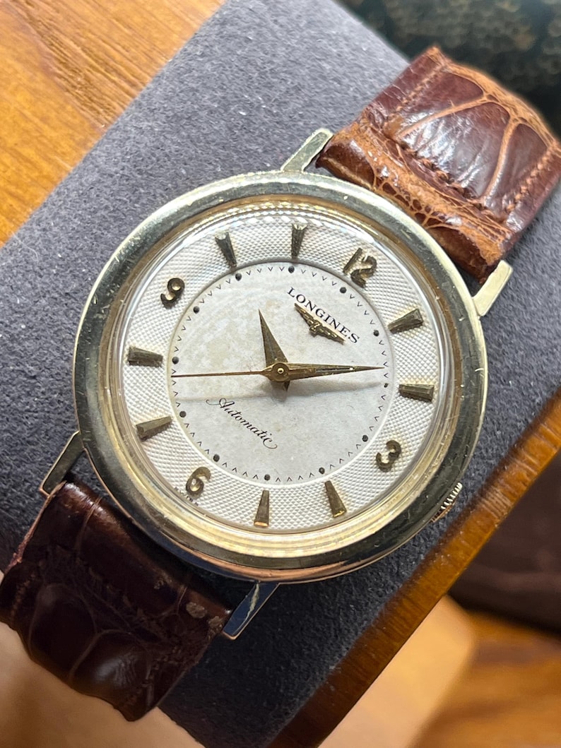 Puede incluir: Un reloj de pulsera autom&aacute;tico Longines vintage con esfera plateada, agujas e indicadores dorados y correa de cuero marr&oacute;n. La esfera del reloj muestra el logotipo de Longines y la palabra "Automatic". El reloj tiene un bisel dorado.