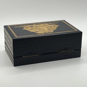 Puede incluir: Una caja rectangular negra con un dise&ntilde;o de barco dorado en la tapa. La caja tiene detalles dorados en los laterales y una apertura con bisagras. Probablemente para guardar joyas o peque&ntilde;os recuerdos.