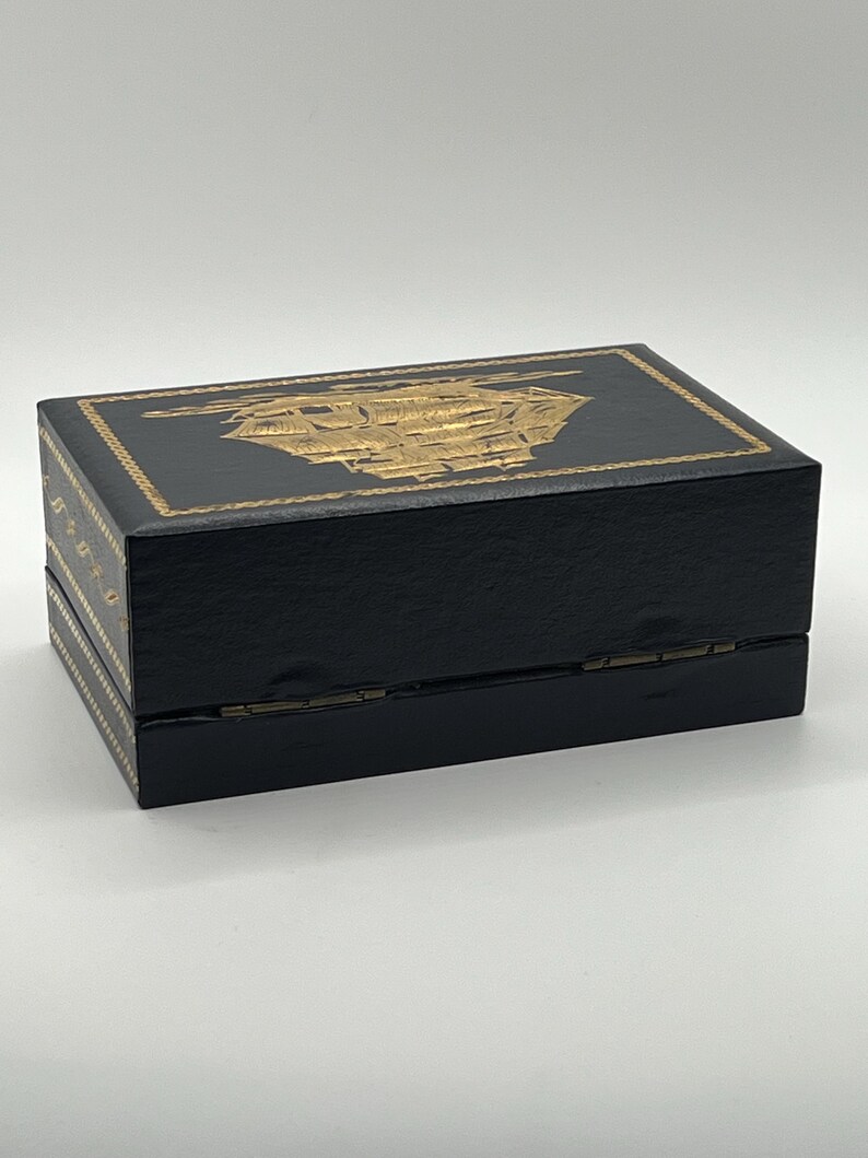 Puede incluir: Una caja rectangular negra con un dise&ntilde;o de barco dorado en la tapa. La caja tiene detalles dorados en los laterales y una apertura con bisagras. Probablemente para guardar joyas o peque&ntilde;os recuerdos.