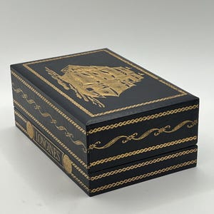 Puede incluir: Caja rectangular negra con detalles dorados. La parte superior presenta una ilustraci&oacute;n dorada de un velero. La caja tiene adornos dorados y la palabra "LONGINES" es visible. Probablemente una caja de reloj.