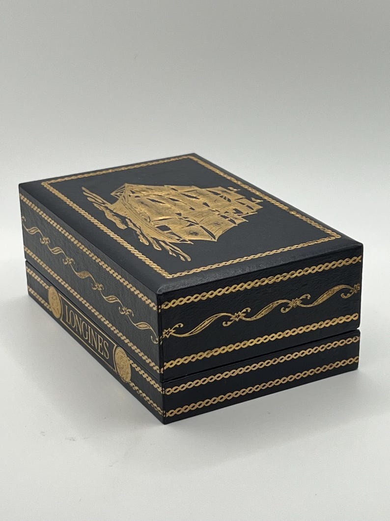 Puede incluir: Caja rectangular negra con detalles dorados. La parte superior presenta una ilustraci&oacute;n dorada de un velero. La caja tiene adornos dorados y la palabra "LONGINES" es visible. Probablemente una caja de reloj.