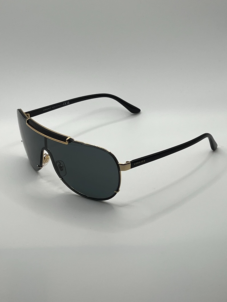 Peut inclure: Lunettes de soleil noires et dor&eacute;es avec un verre en forme d'&eacute;cran. Les lunettes ont une monture noire avec des accents dor&eacute;s et des verres fonc&eacute;s. Le nom de la marque "Versace" est visible sur la branche.