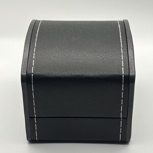 May include: A black, rectangular jewelry box with a textured surface and white stitching along the edges. The box has a curved top and a hinged lid, suggesting it is designed to open and close.