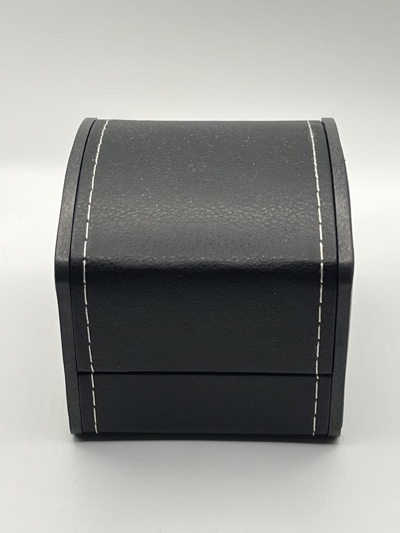 May include: A black, rectangular jewelry box with a textured surface and white stitching along the edges. The box has a curved top and a hinged lid, suggesting it is designed to open and close.